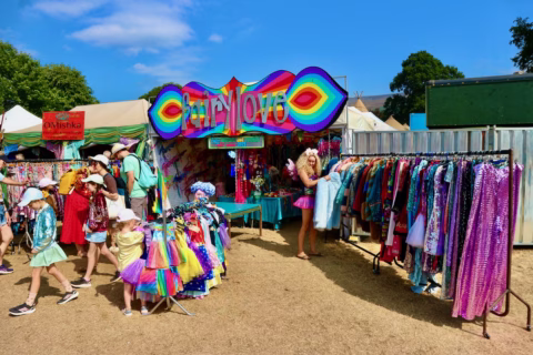 Colourful outdoor market scene with a Fairy Love stall, people in bright costumes and hats, and rails of vibrant clothing under a sunny blue sky. Trees and additional tents are visible in the background.