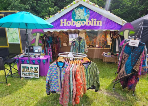 A colourful outdoor market stall with a Hobgoblin sign sells patterned jackets, ponchos, and accessories. An umbrella shades a Face Painting table. The scene is festive and vibrant.