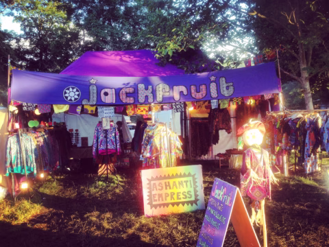 Outdoor market stall with a purple Jackfruit sign, colourful clothing on racks, a mannequin in a dress, and an Ashanti Empress sign, with trees and sunlight in the background.