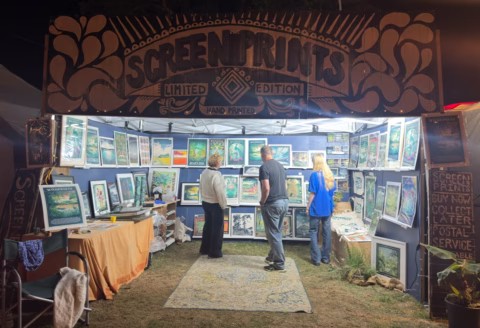 Three people view colourful art prints in a festive stall labelled Screenprints Limited Edition. The stall displays numerous framed prints, and the area is warmly lit, creating an inviting atmosphere for visitors.