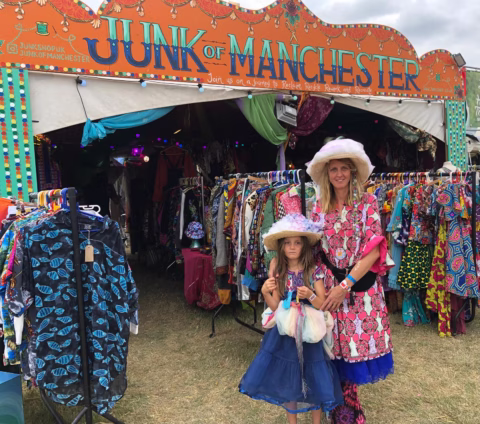 Two people in colourful clothes and hats stand in front of a bright, patterned tent labelled Junk of Manchester, with racks of vibrant clothing visible inside and outside the tent at an outdoor event.