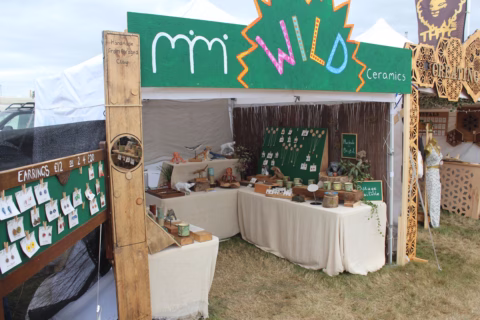 A market stall with a green sign reading WILD Ceramics displays handmade ceramics and jewellery on tables and boards, set up outdoors on grass under a white marquee.