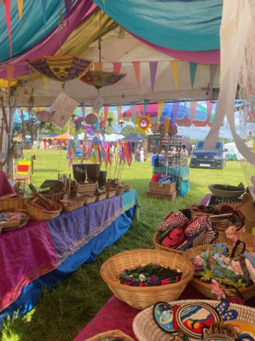 A colourful outdoor market stall displays woven baskets, beaded jewellery, drums, and vibrant crafts under a decorated tent with flags and hanging ornaments. People and a car are visible outside on a grassy field.