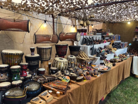 A market stall displays various traditional musical instruments, including drums, percussion instruments, and shakers, arranged on tables covered with cloth under camo netting.