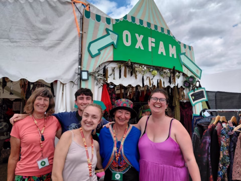 Five smiling people stand in front of an Oxfam tent at a festival, wearing colourful outfits and badges. The tent is decorated with clothes and has a green Oxfam sign above the entrance.