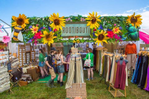 Colourful outdoor boutique with large faux sunflowers, dresses on rails, jewellery displays, and people browsing. The sign above reads ROOTIBOUTI, decorated with flowers, bees, and greenery. Grass and blue sky visible.