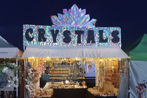 A market stall at night with a colourful, crystal-themed sign reading CRYSTALS, decorated with lights and selling various crystal and jewellery items.