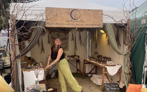 A smiling woman stands in front of a stand labelled earth remedies, displaying bottles and products on tables, with decorative branches and natural-themed decor in the background.
