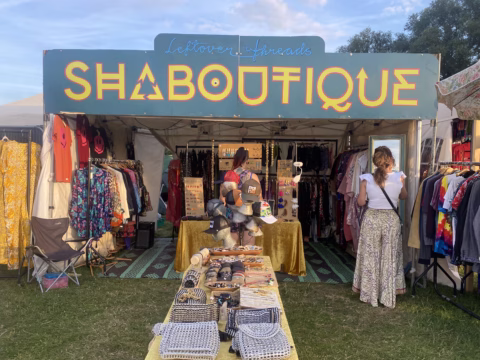 Outdoor vintage clothing boutique with colourful dresses and accessories on rails, a person sitting behind a table of jewellery, and a customer browsing. Sign reads SHABOUTIQUE..