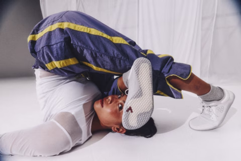 A person wearing a white top, purple trousers with yellow stripes, and white trainers performs a flexible pose, lying on the floor with one leg bent over their head, looking directly at the camera.