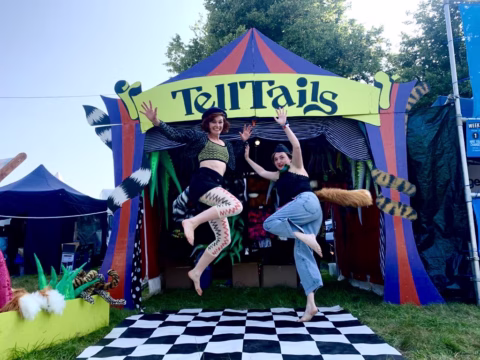 Two people with animal tails jump in front of a colourful tent labelled TellTails at an outdoor event, with a black and white chequered mat on the grass and costume tails displayed around.