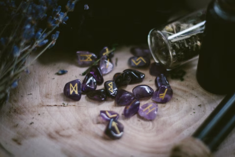 Purple rune stones with gold symbols scattered on a wooden surface, surrounded by a glass vial, a black container, dried herbs, and a wooden wand.