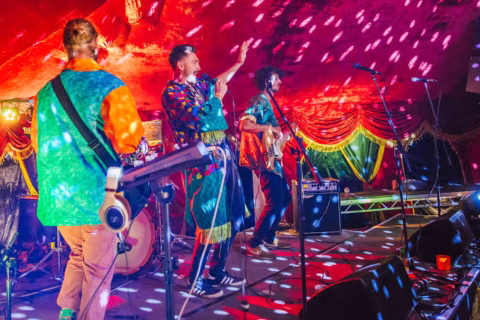 A colourful band performs energetically on stage under vibrant, disco-style lighting, surrounded by musical instruments and decorative drapes.