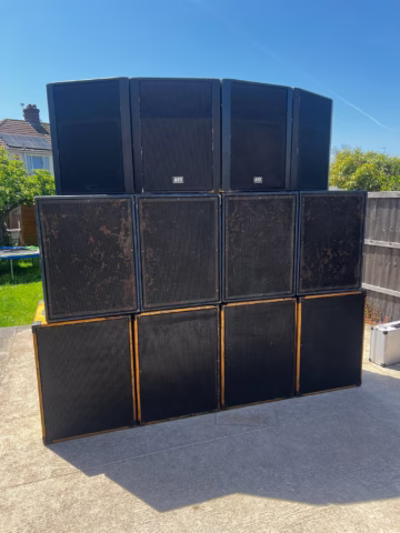 A large stack of black speakers is set up outdoors on a sunny day, arranged in three rows on a concrete surface, with a garden and wooden fence in the background.