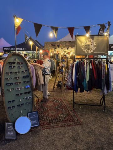 A vintage clothing stall at an outdoor market at dusk, featuring rails of colourful jackets, sunglasses on display, rugs, and fairy lights overhead. A person browses through clothes under a blue evening sky.