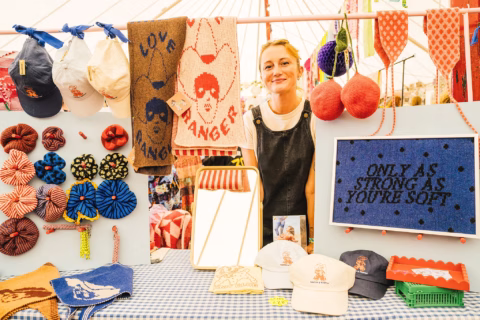 A smiling vendor stands behind a stall displaying hats, knitted items, and a sign reading ONLY AS STRONG AS YOU'RE SOFT at a colourful market stall.