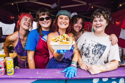 Five smiling people stand at a colourful food stall, one holding a tray of loaded chips towards the camera. Bright drinks are visible to the side, and fairy lights hang above them.