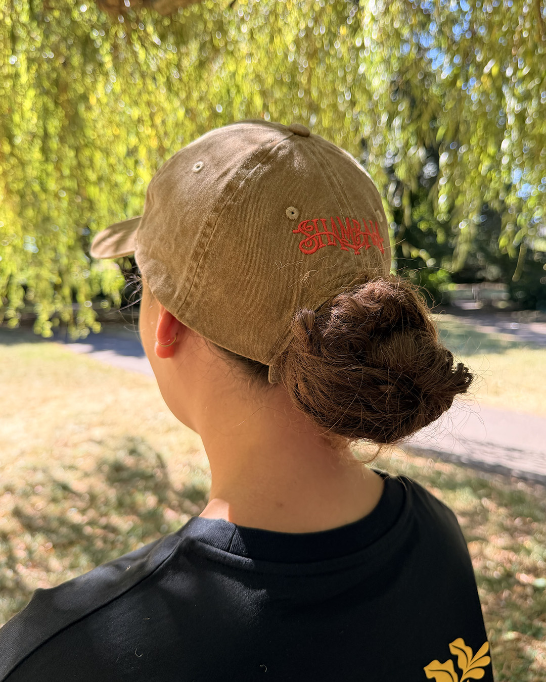 A person with long brown hair in a low bun is wearing the SHAMBALA BASEBALL CAP and a black shirt whilst standing outdoors under leafy green trees on a sunny day.