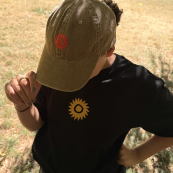 A person stands outdoors on dry grass, holding the brim of the SHAMBALA BASEBALL CAP, which is khaki with a red sun symbol, and wears a black shirt featuring a yellow sun symbol.