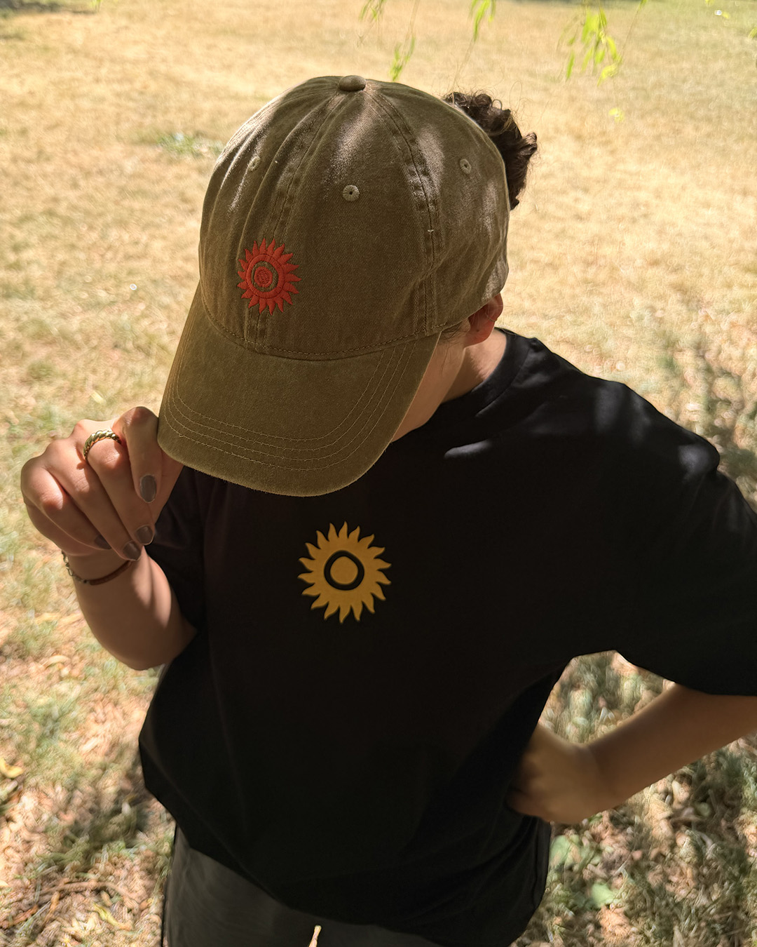 A person stands outdoors on dry grass, holding the brim of the SHAMBALA BASEBALL CAP, which is khaki with a red sun symbol, and wears a black shirt featuring a yellow sun symbol.