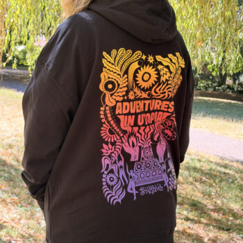 A person with long hair wears the HOODIE: SHAMBALA x SOPHIE BASS, featuring a colourful psychedelic design and the words “Adventures in Utopia” on the back, whilst standing outdoors near grass and trees.