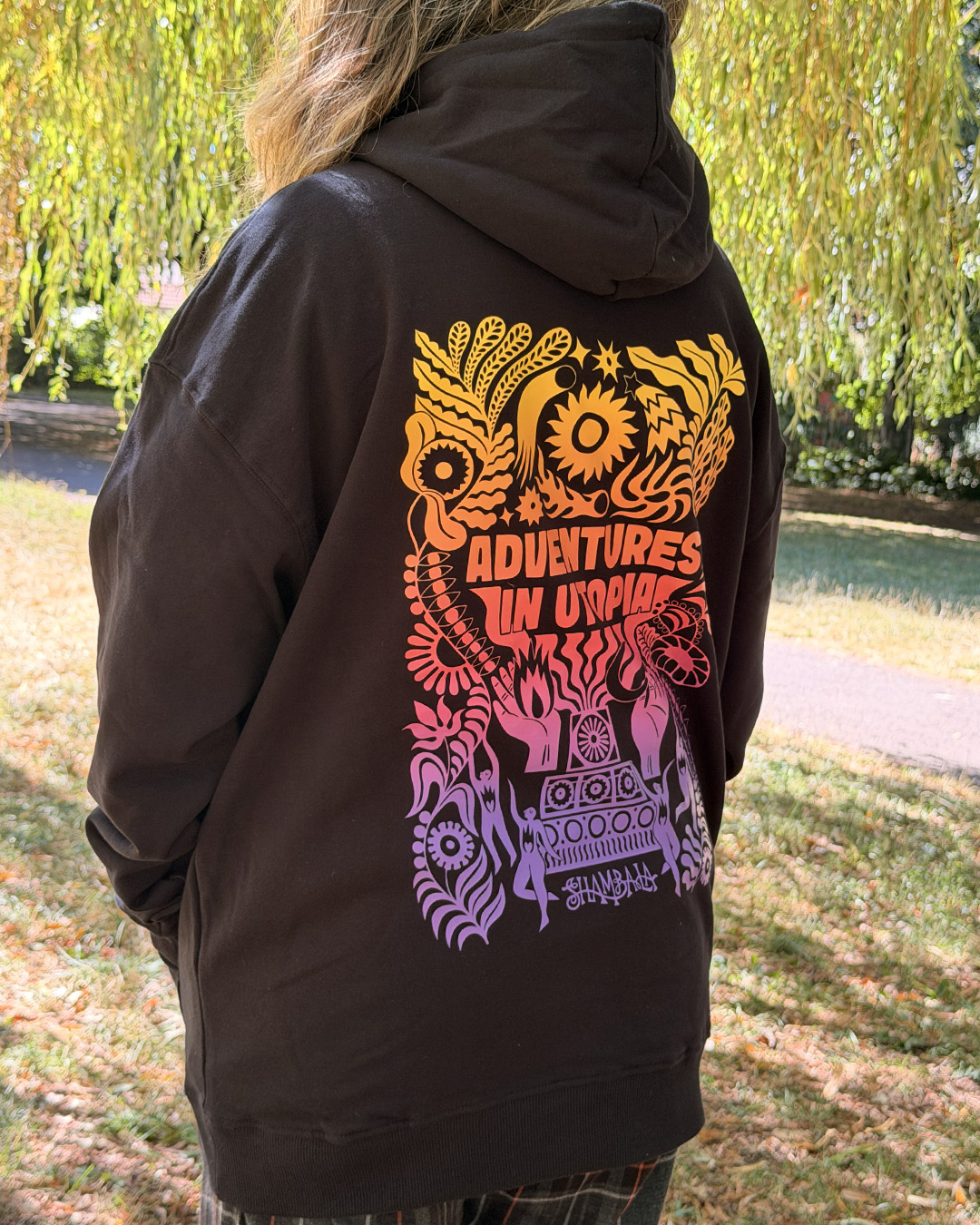 A person with long hair wears the HOODIE: SHAMBALA x SOPHIE BASS, featuring a colourful psychedelic design and the words “Adventures in Utopia” on the back, whilst standing outdoors near grass and trees.