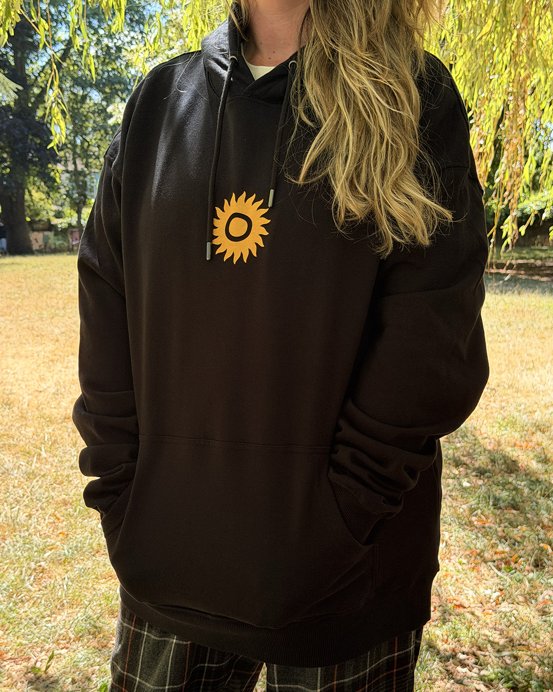 A person with long, wavy blonde hair wears the HOODIE: SHAMBALA x SOPHIE BASS, featuring a yellow sunburst design on the chest, whilst standing outdoors on grass under sunlight with trees in the background.