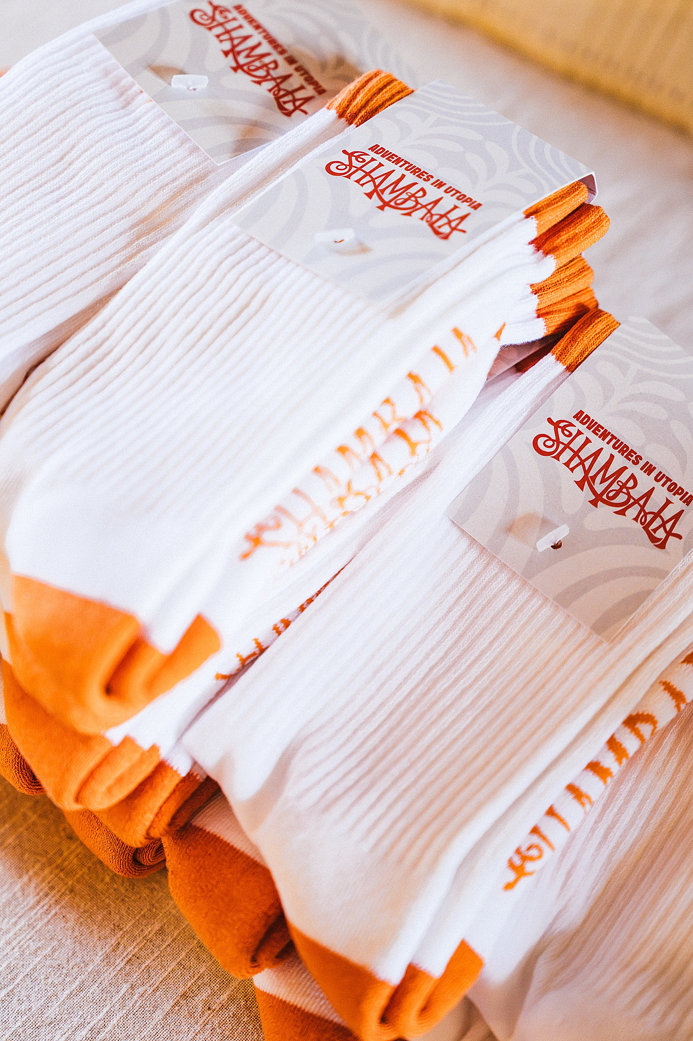 A stack of white SHAMBALA CREW SPORT SOCKS featuring orange toes and heels, each pair wrapped with a label that reads “Adventures in Utopia SHAMBALA” in orange text.