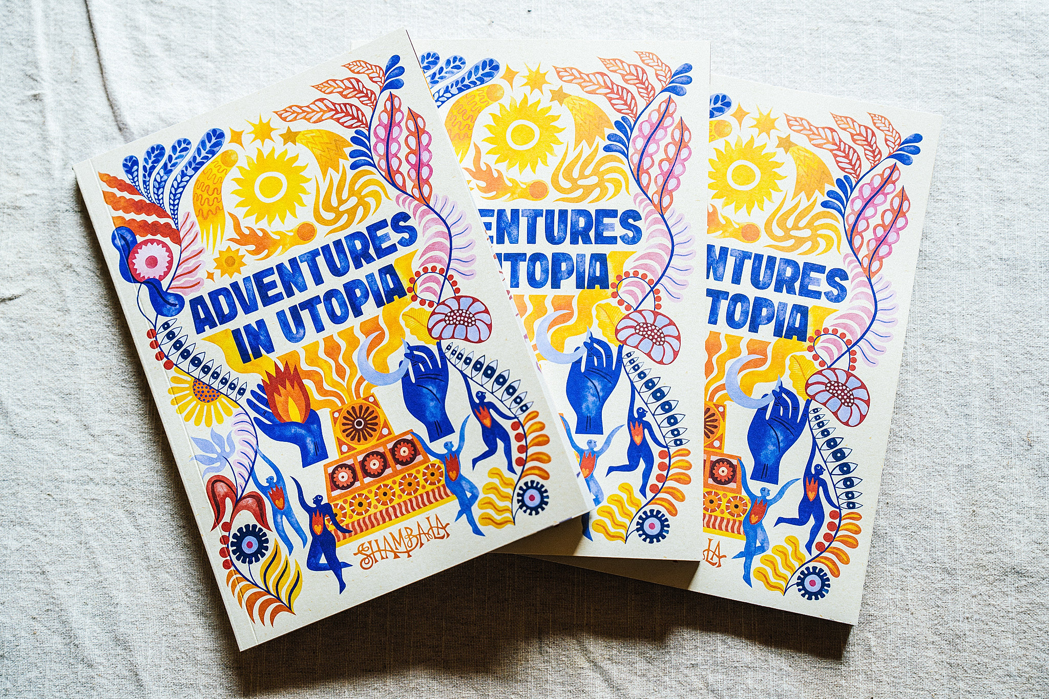 Three vibrant SHAMBALA 2026 PRINTED PROGRAMME booklets, featuring abstract floral and hand-drawn designs in blue, yellow, and orange, displayed on a light fabric background.