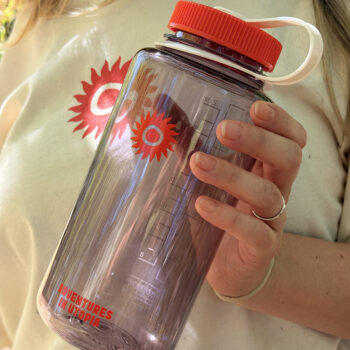 A person wearing a cream shirt with a red sunburst design holds the 1L WATER BOTTLE: SHAMBALA x NALGENE, a large translucent bottle featuring matching red sunburst art, a red lid with a white handle, and text that reads ADVENTURES IN UTOPIA.