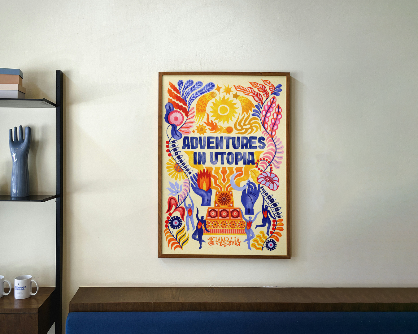 A LIMITED EDITION A2 ART PRINT: SHAMBALA x SOPHIE BASS hangs on the wall, featuring “ADVENTURES IN UTOPIA” amid blue, orange, and yellow abstract shapes, plants, sun, hands, and faces. Books and a decorative hand rest on a nearby shelf.