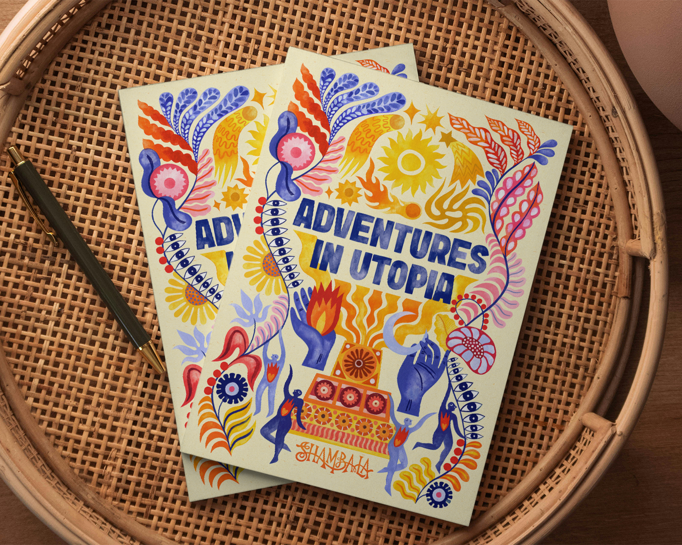 Two SHAMBALA 2026 PRINTED PROGRAMME booklets with vibrant, abstract floral and flame covers are placed on a woven round tray next to a black pen.