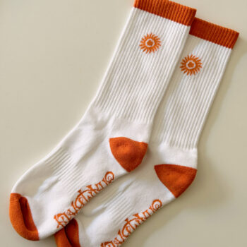 A pair of SHAMBALA CREW SPORT SOCKS in white with orange toes, heels, and cuffs, featuring a sun motif near the top and orange lettering along the sole, displayed side by side on a neutral background.