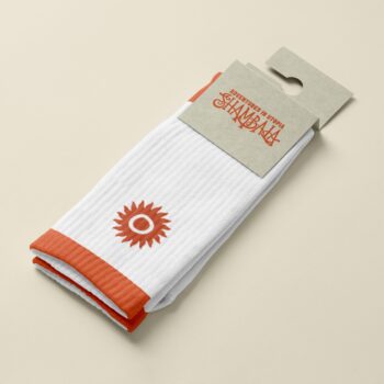 The SHAMBALA CREW SPORT SOCKS are white with orange toes, heels, and cuffs, featuring an orange sun symbol near the ankle. They come packaged with a brown cardboard label on a beige background.