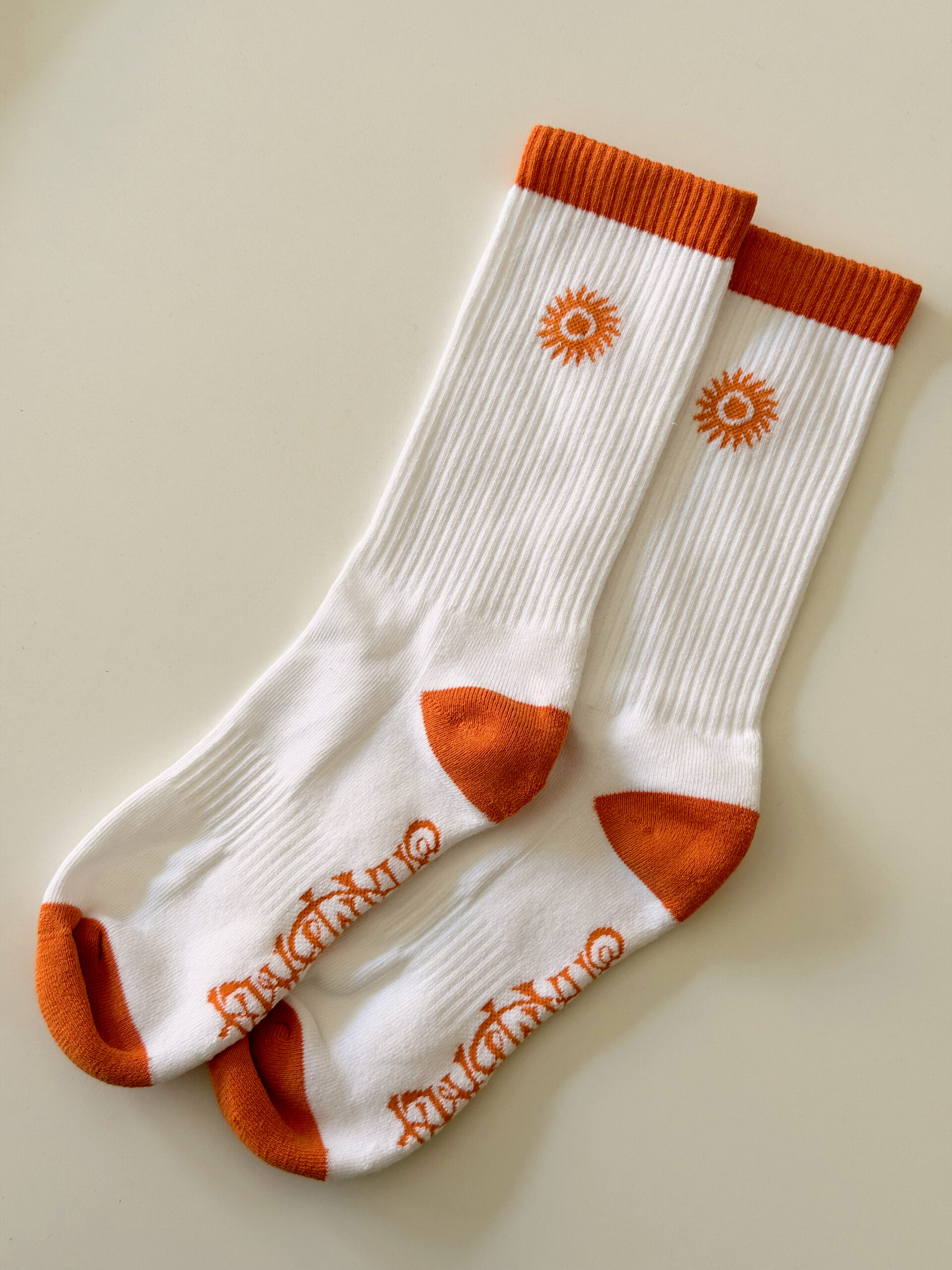 A pair of SHAMBALA CREW SPORT SOCKS in white with orange toes, heels, and cuffs, featuring a sun motif near the top and orange lettering along the sole, displayed side by side on a neutral background.