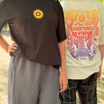 Two people stand outdoors. One faces forward, wearing a black T-SHIRT: SHAMBALA x SOPHIE BASS featuring a yellow sun symbol. The other has their back turned, wearing a white T-SHIRT: SHAMBALA x SOPHIE BASS with colourful designs and “ADVENTURES IN UTOPIA” printed on the back.
