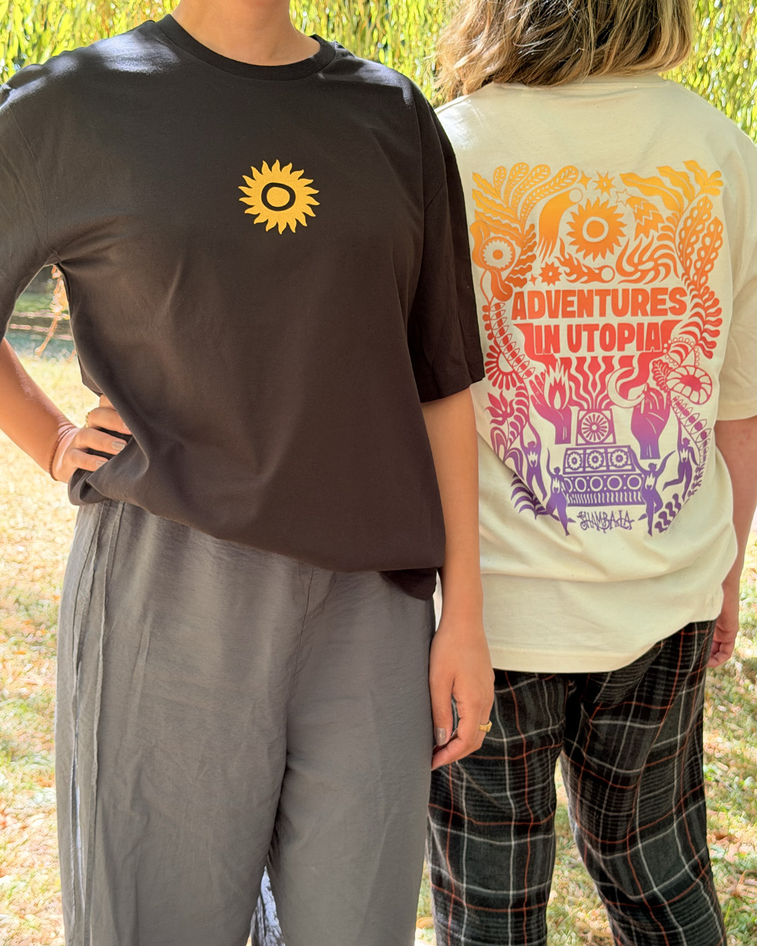 Two people stand outdoors. One faces forward, wearing a black T-SHIRT: SHAMBALA x SOPHIE BASS featuring a yellow sun symbol. The other has their back turned, wearing a white T-SHIRT: SHAMBALA x SOPHIE BASS with colourful designs and “ADVENTURES IN UTOPIA” printed on the back.