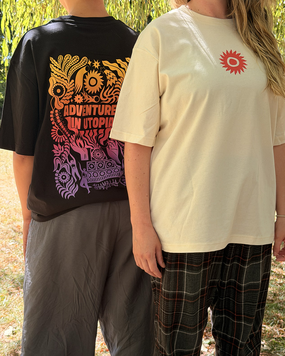 Two people stand side by side outdoors. One wears the T-SHIRT: SHAMBALA x SOPHIE BASS in black, featuring colourful designs and “ADVENTURE IN UTOPIA” text on the back; the other wears a light-coloured T-shirt with a small red sun graphic on the front.
