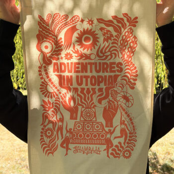 A person holds the TEA TOWEL: SHAMBALA x SOPHIE BASS, featuring a bold orange “Adventures in Utopia” design with whimsical patterns, plants, and a central sun motif on beige cloth. The scene is set outdoors amidst greenery and sunlight.