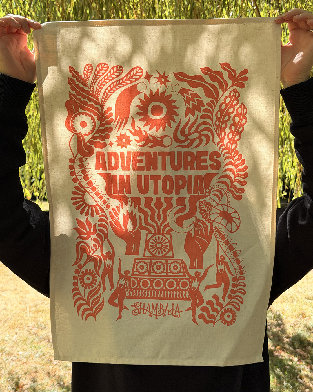 A person holds the TEA TOWEL: SHAMBALA x SOPHIE BASS, featuring a bold orange “Adventures in Utopia” design with whimsical patterns, plants, and a central sun motif on beige cloth. The scene is set outdoors amidst greenery and sunlight.