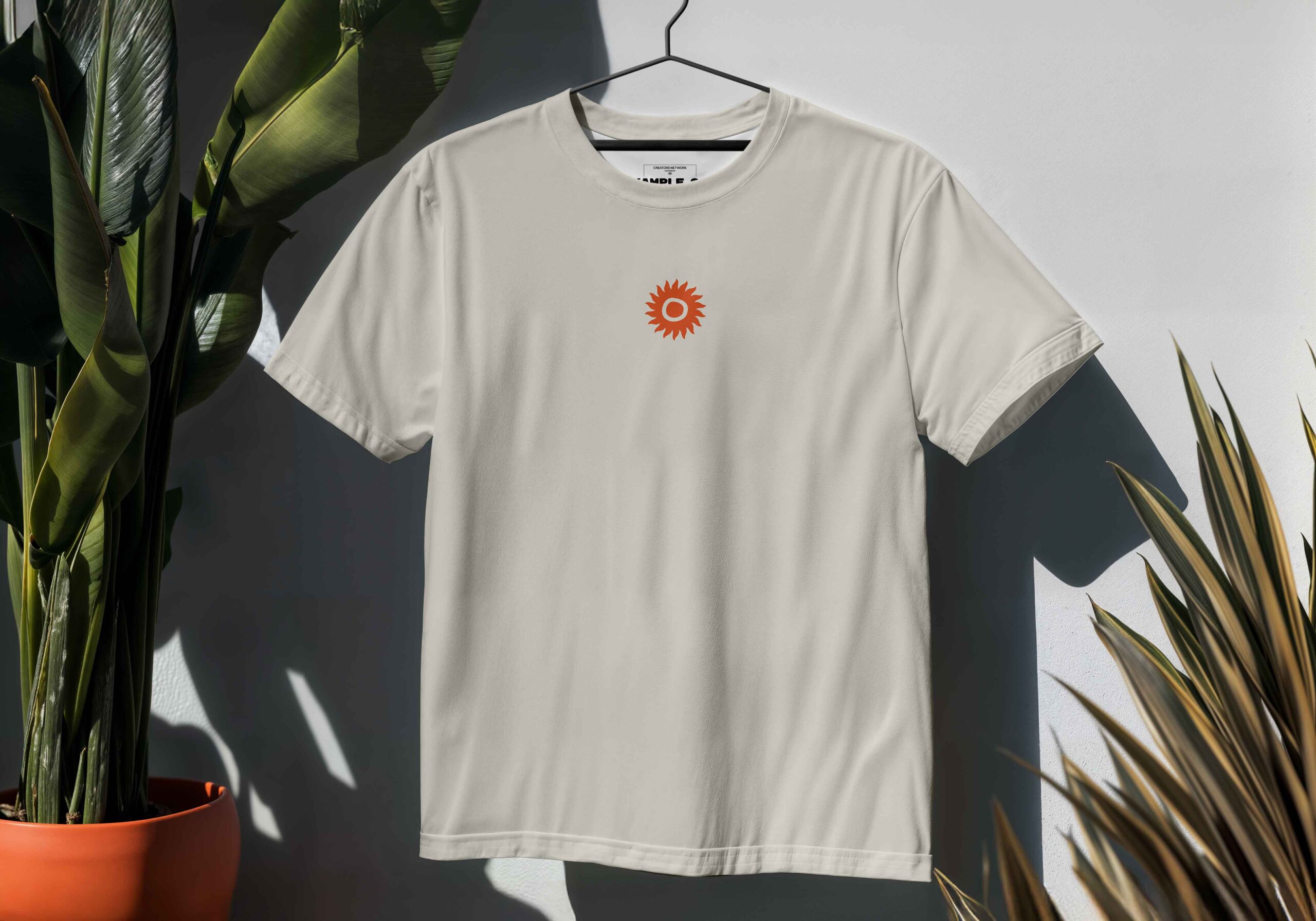 The T-SHIRT: SHAMBALA x SOPHIE BASS, featuring a small red sun on the chest, hangs on a hanger among green potted plants with sunlight creating shadows on the wall.