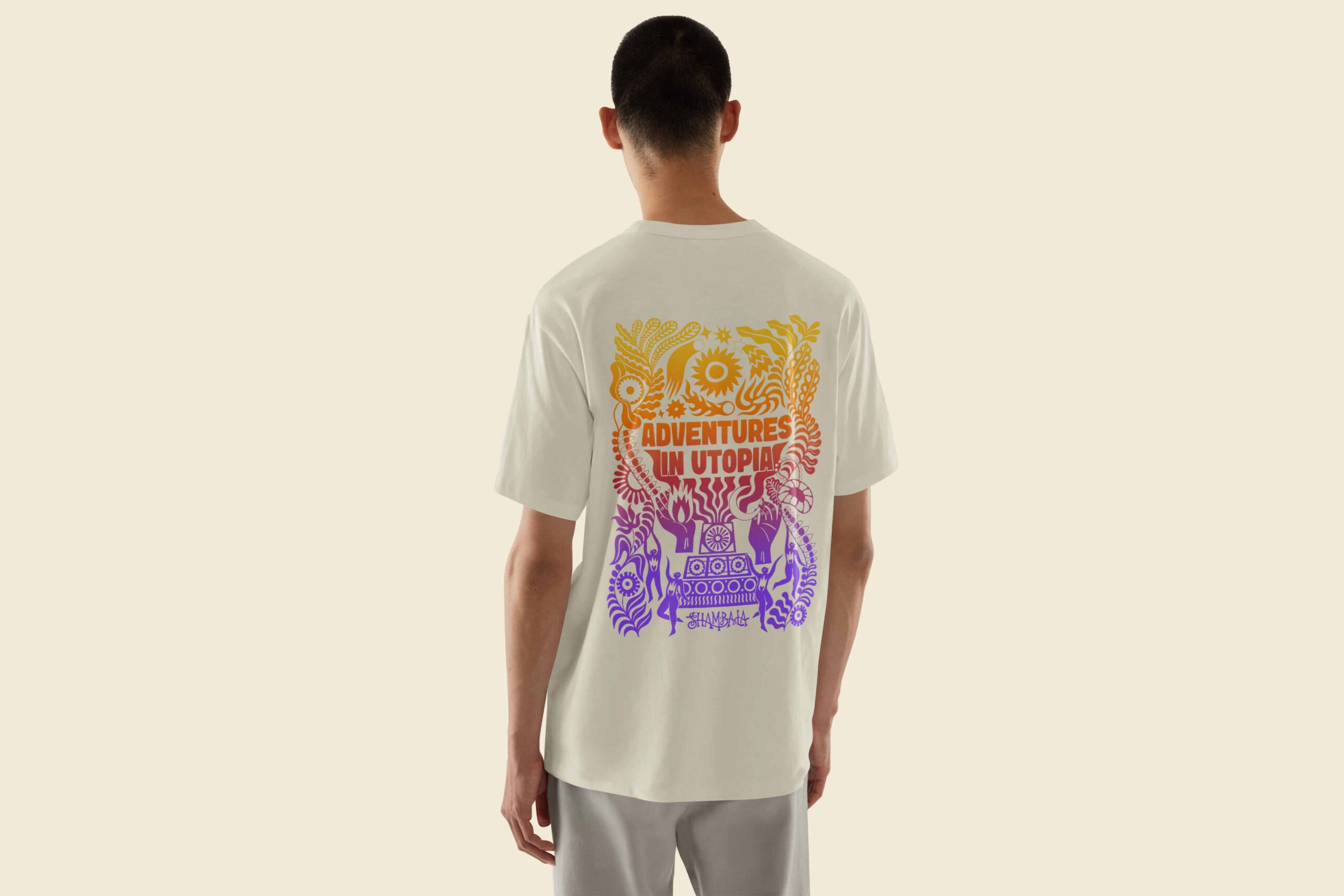 A person is seen from behind wearing the T-SHIRT: SHAMBALA x SOPHIE BASS, a white tee with colourful abstract artwork and "Adventures in Utopia" text, featuring orange, pink, and purple shapes and patterns.