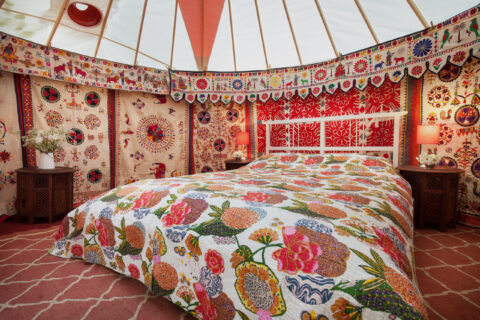 A cosy yurt interior with colourful floral bedding, vibrant patterned wall hangings, two wooden bedside tables with lamps, and a vase of flowers, creating a warm and inviting atmosphere.