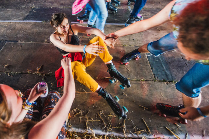 A young woman in yellow trousers laughs as she falls on roller skates, reaching for help from friends, surrounded by people skating on a wooden floor.