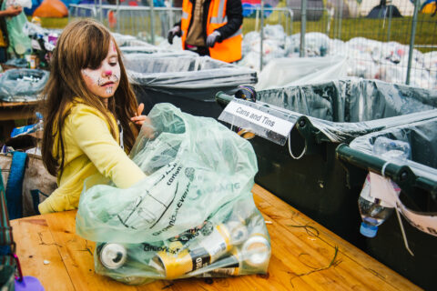 A young girl with face paint sorts empty cans into a recycling bin labelled “Cans and Tins,” surrounded by bags of recyclables—capturing the eco-friendly spirit rooted in Shambala's Festival history.