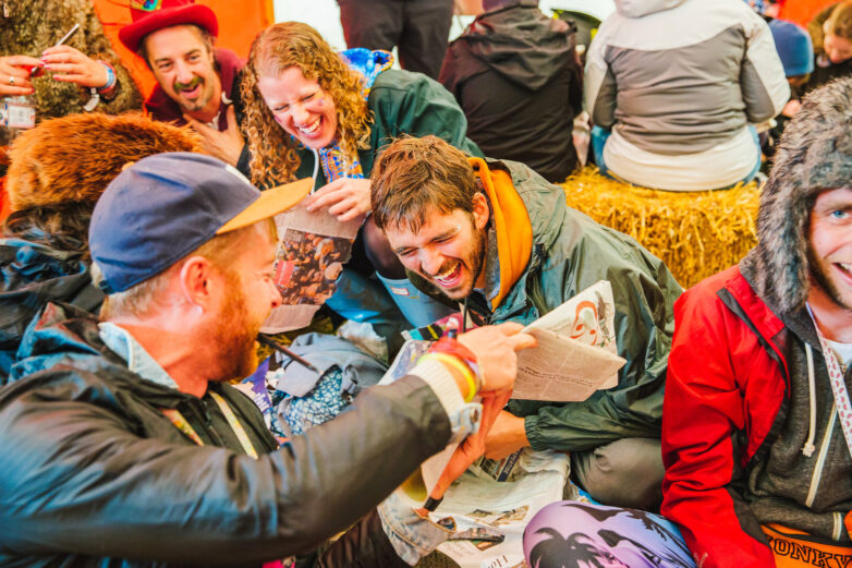 A group of people in colourful, casual clothes sit closely together on hay bales, laughing and reading newspapers.