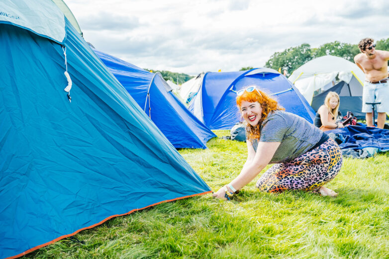 A smiling woman crouches as she secures a tent at a campsite surrounded by other. The weather looks sunny with scattered clouds.