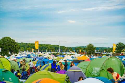 A colourful outdoor Shambala scene with many tents set up on grassy ground, people walking and talking among them, and yellow flags on poles. Trees and a blue sky are visible in the background.