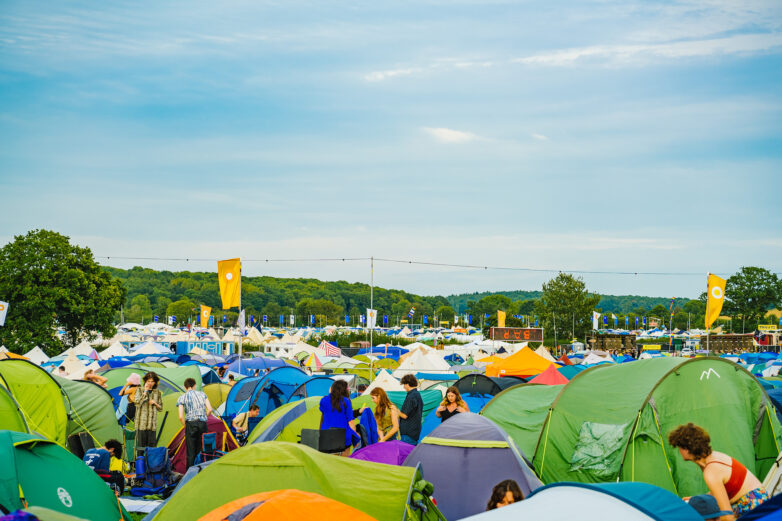A colourful outdoor Shambala scene with many tents set up on grassy ground, people walking and talking among them, and yellow flags on poles. Trees and a blue sky are visible in the background.