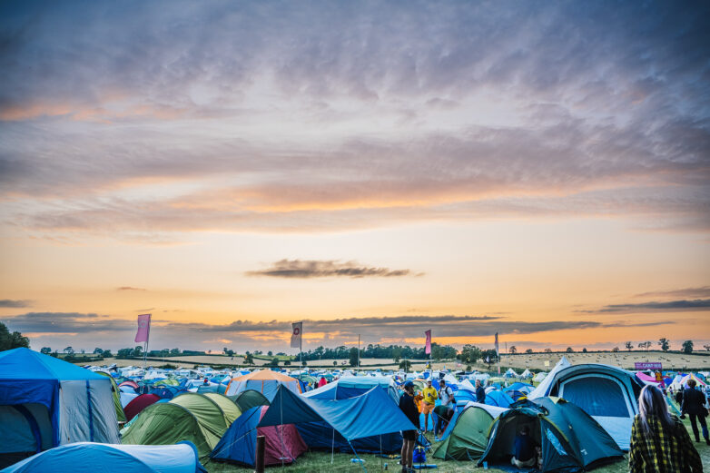 Rows of colourful tents set up on a grassy field at sunset, with a dramatic sky overhead and people walking among the tents; festival flags and trees are visible in the background.